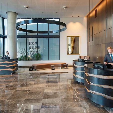 Hotel Pullman Brisbane Airport Exteriér fotografie The image shows a modern hotel lobby. There are two reception desks made of dark-colored materials, and staff members are attending to guests at these desks. The lobby features large windows that allow natural light to fill the space, and there are p