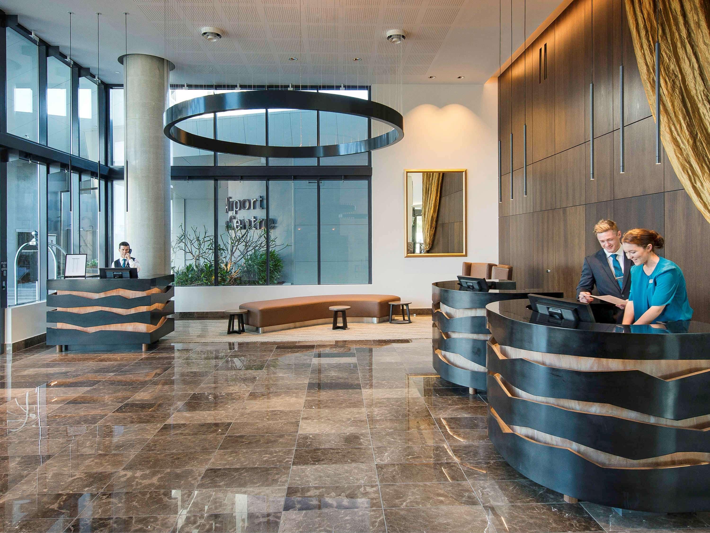 Hotel Pullman Brisbane Airport Exteriér fotografie The image shows a modern hotel lobby. There are two reception desks made of dark-colored materials, and staff members are attending to guests at these desks. The lobby features large windows that allow natural light to fill the space, and there are p